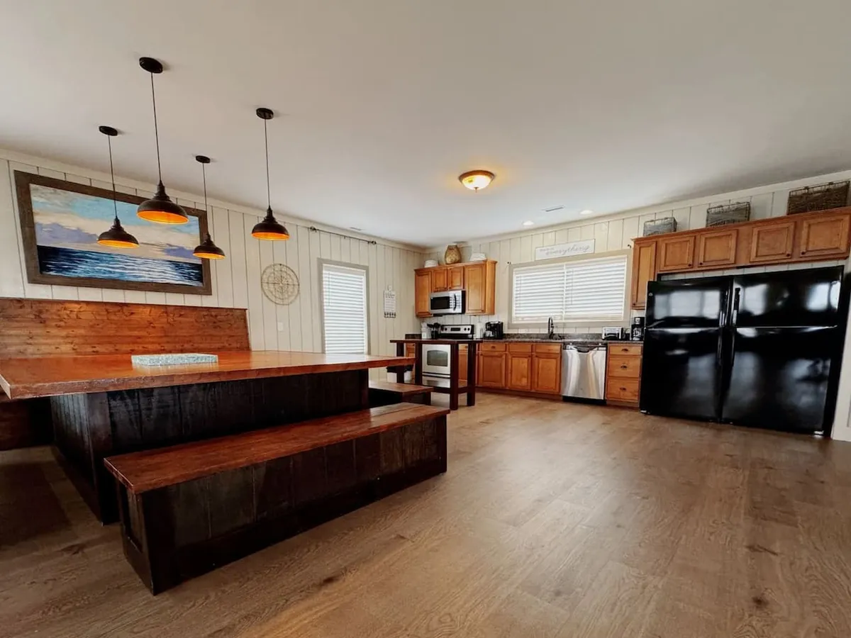 A view of a substantial wooden dining table positioned near sleek black appliances and under elegant pendant lights. The arrangement emphasizes togetherness and the functionality of the kitchen for group cooking and dining.