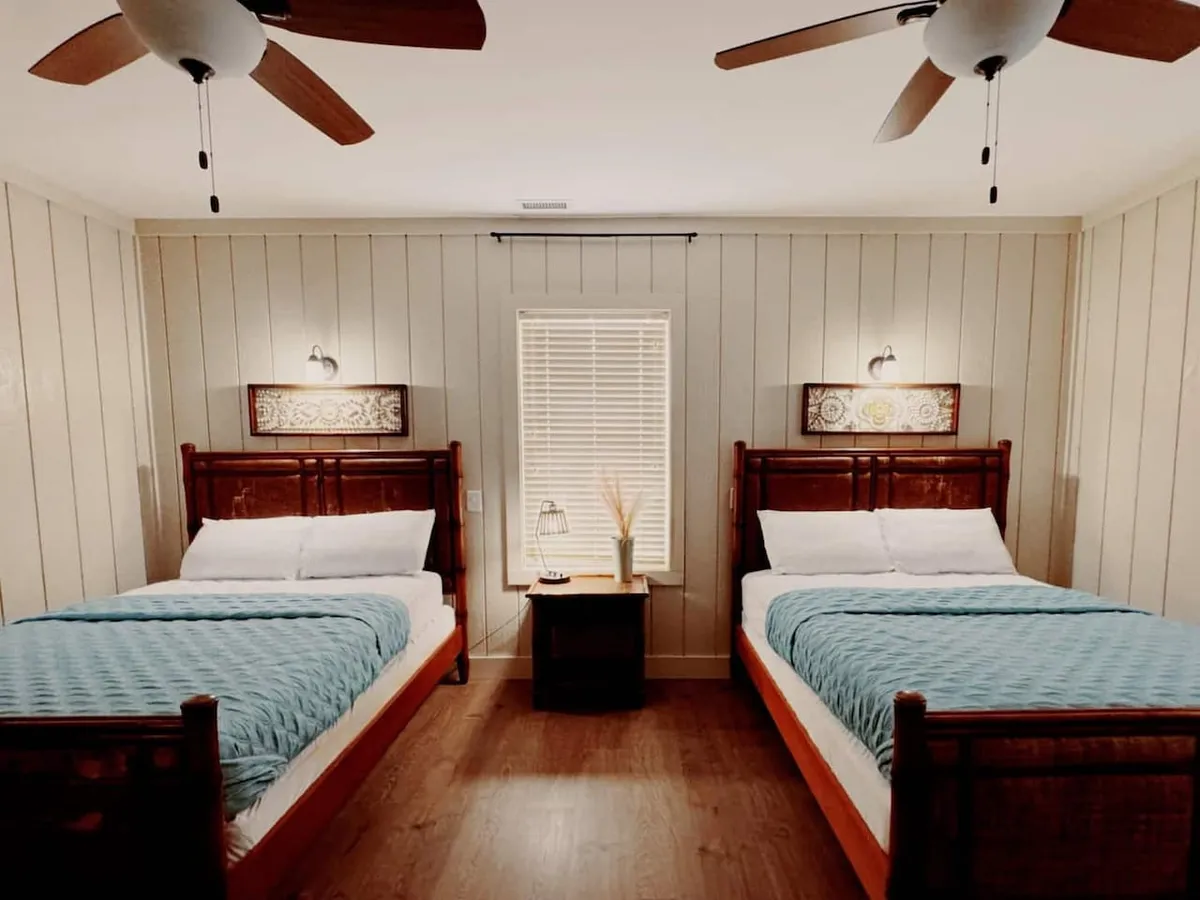 A serene bedroom featuring two wooden beds with plush bedding, a nightstand with a lamp ideal for reading, stylish decorative wall art, and a window with blinds letting in natural light. The layout and cozy decor create a welcoming and restful atmosphere, perfect for winding down after a long day.
