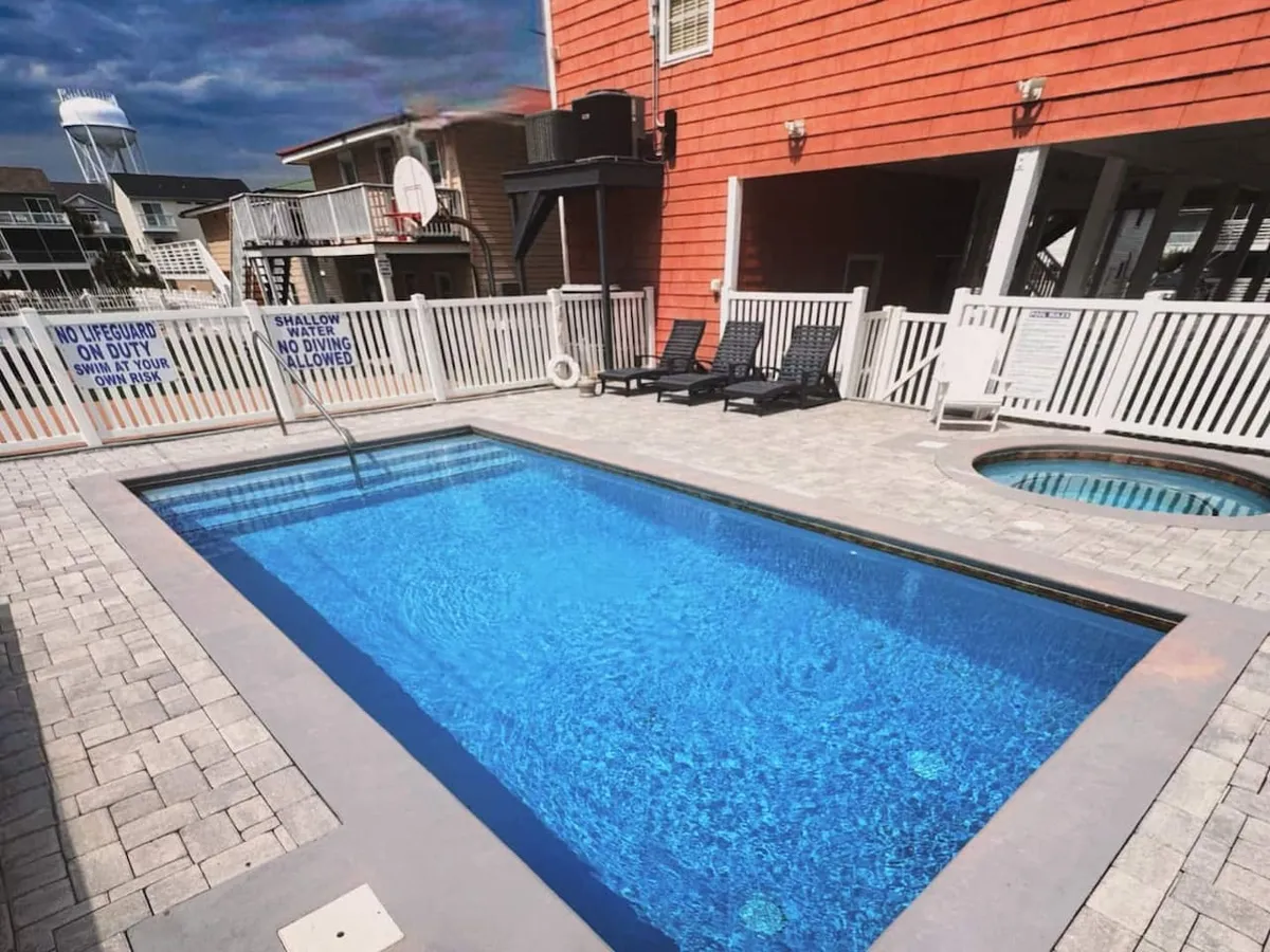 A fenced backyard scene showing a family-friendly swimming pool with an attached hot tub, surrounded by lounge chairs and visible safety signs. The fence ensures a secure environment for both children and pets, while the layout invites casual poolside relaxation and socializing.