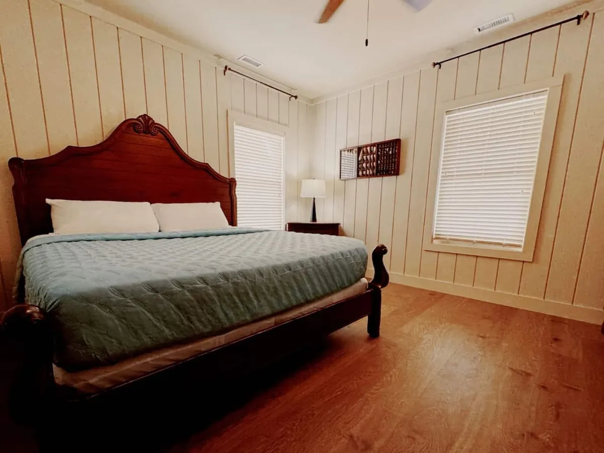 A tranquil bedroom scene with a comfortable bed, a softly glowing lamp on the bedside table, and windows that allow in plenty of natural light. The arrangement is simple and calming, emphasizing the promise of a peaceful nap or quiet reading time.