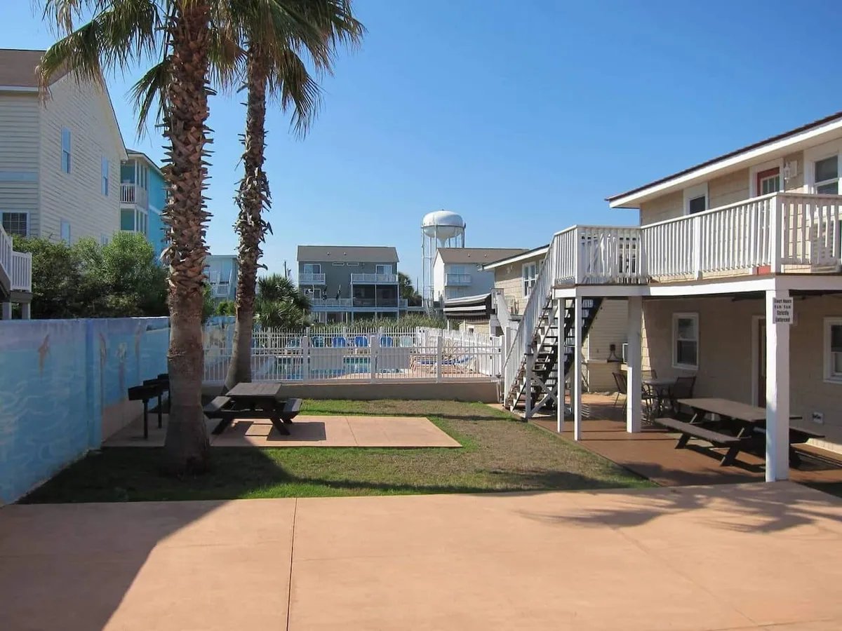 An inviting exterior shot featuring a swimming pool bordered by picnic tables, tall palm trees, and a staircase leading up to the home’s upper level. The setting is perfect for poolside meals, sunbathing, and leisurely mornings under the palm trees.