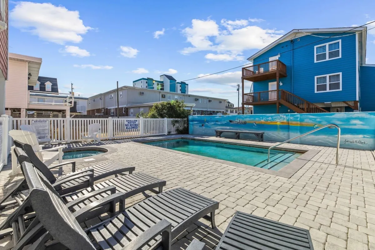 A bright exterior scene featuring a sparkling swimming pool with an attached hot tub, surrounded by lounge chairs and a striking colorful mural on the wall. The area is designed for both relaxation and social gatherings, with ample space for sunbathing and cheerful group activities.