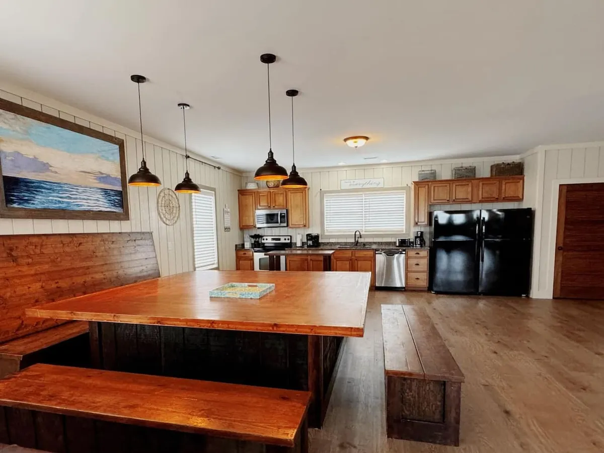 This image features a large wooden dining table under pendant lights, with stylish kitchen cabinetry and artwork adorning the wall. The setting is warm and inviting, emphasizing a space designed for both cooking and communal meals.