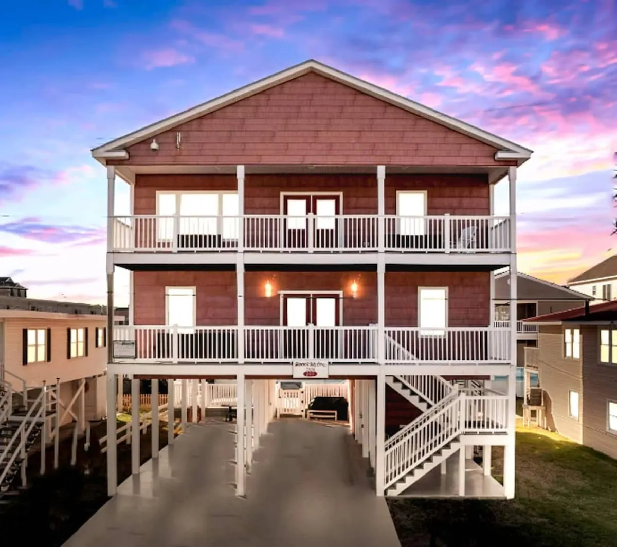 A vibrant exterior view of a three-story vacation home featuring white railings on spacious balconies and a staircase leading up to the entrance. The scene radiates energy, suggesting gatherings or relaxation on the balconies at sunset after a fun night out.