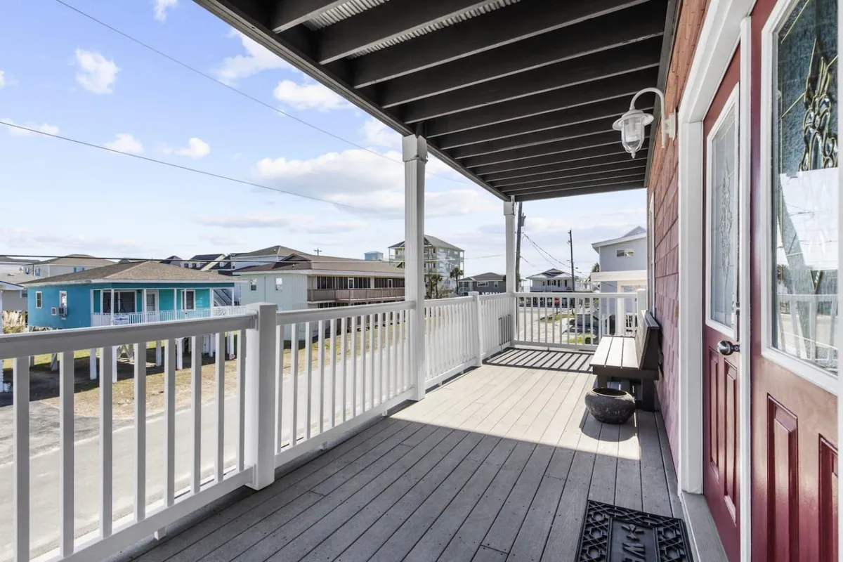 An exterior view of a porch with inviting outdoor seating and a railing, looking out toward neighboring houses. The space is perfect for enjoying meals outside with fresh air and scenic views.