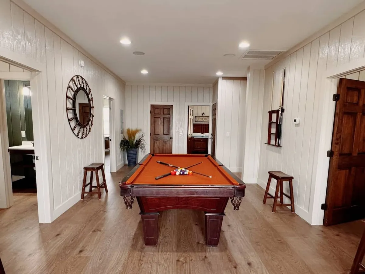 An inviting game room centers around a pool table, flanked by wooden stools and a decorative mirror. Double wooden doors add warmth, while the overall setup encourages friendly competition and social interaction. The lighting and casual arrangement create a fun, laid-back environment.