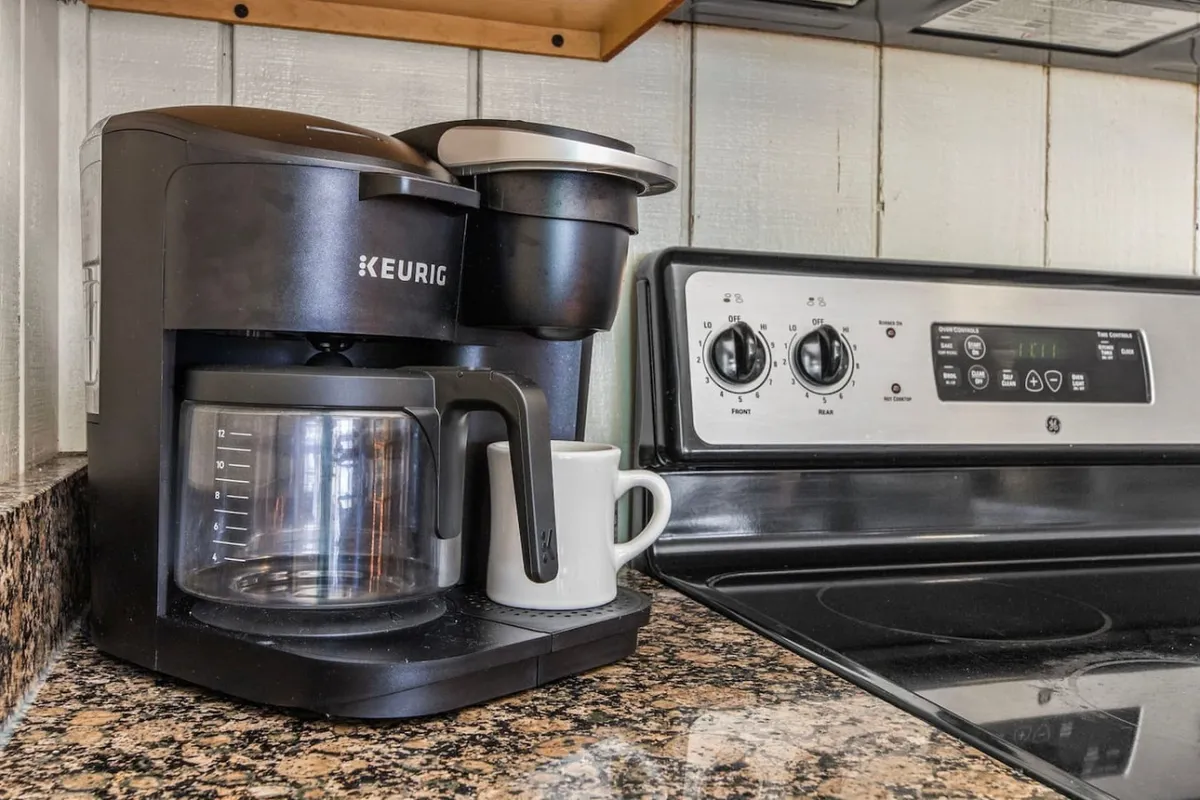 A close-up of a kitchen corner featuring a Keurig coffee maker, glass carafe, white coffee cup, and stainless steel stove. The scene feels warm and homey, suggesting the start of a day of exploration or the perfect finish to an evening in.