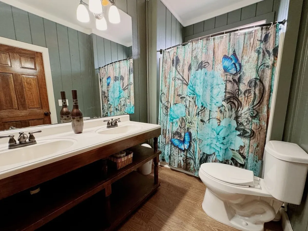 A modern bathroom with a double sink vanity, a floral shower curtain, and a wooden door. The bright, clean design evokes a spa-like atmosphere—ideal for morning routines or relaxing after a day of adventure.