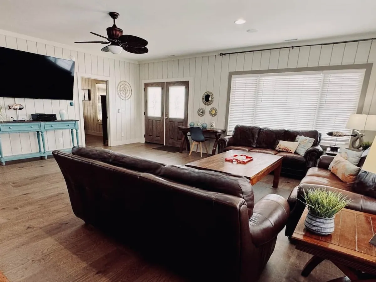 A spacious, sunlit living room features a large leather sofa and wood coffee table, ideal for lounging and gathering. The blue console table and double entry doors provide a welcoming entry point, while artwork on the walls adds a personal, homey touch. The arrangement suggests ample seating for group relaxation and social activities, with an inviting, comfortable atmosphere.