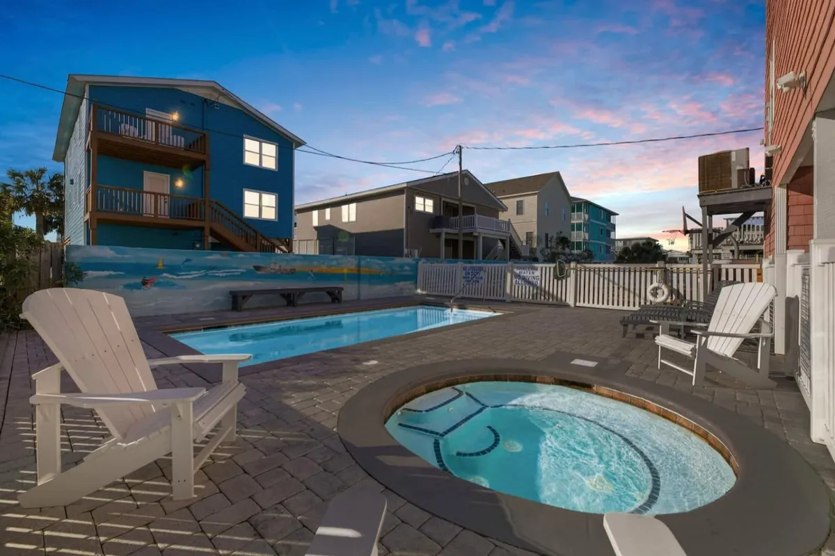 A cheerful, fenced backyard featuring a sparkling swimming pool, a hot tub, colorful houses in the background, and Adirondack chairs for relaxing. The space invites group activities such as swimming, sunbathing, and socializing in a secure, private setting.