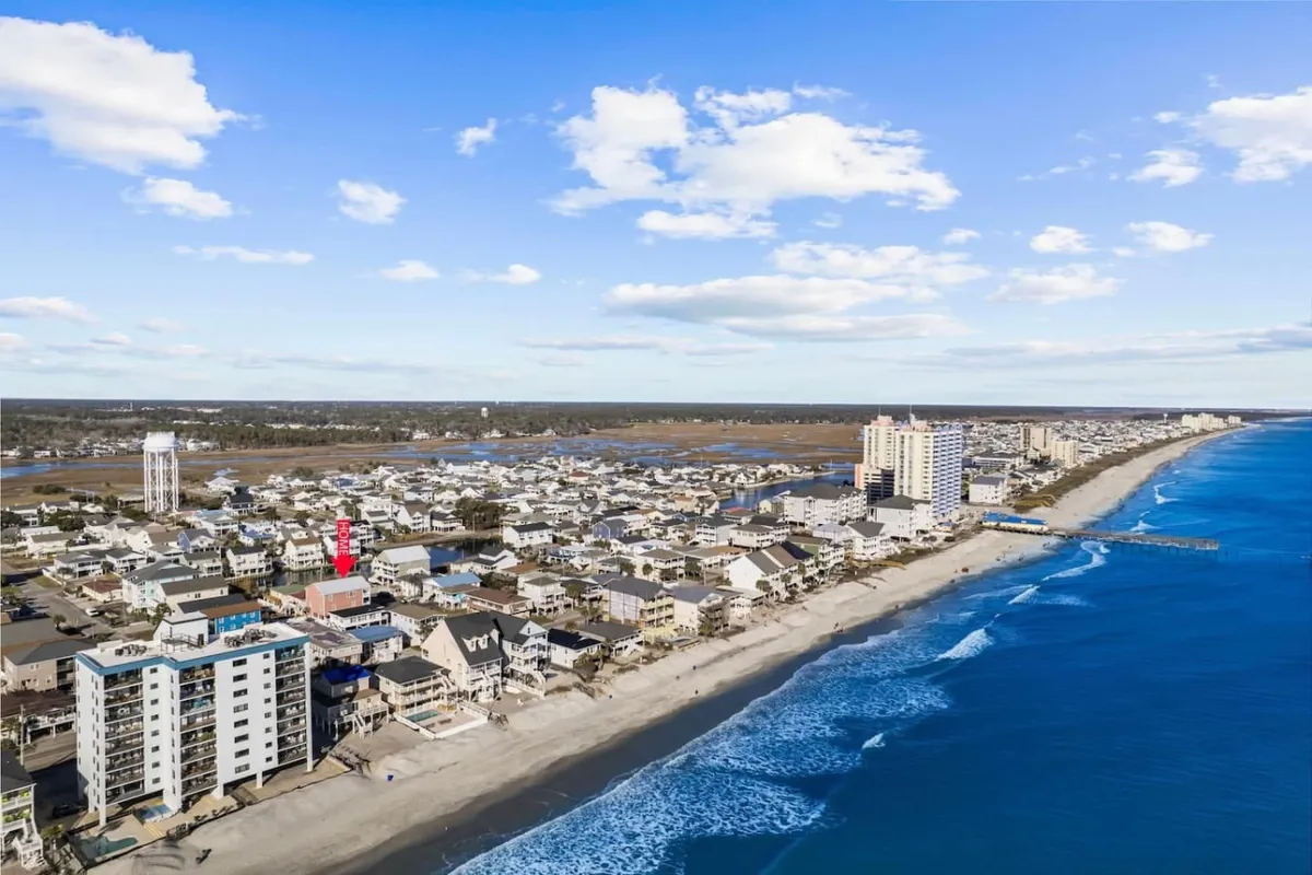 A sweeping view of Cherry Grove Beach with beachfront properties, the ocean, a boardwalk, pier, and the town’s buildings in the background. The scene is lively with people swimming, sunbathing, and exploring the local coastal attractions, evoking a vibrant, family-friendly atmosphere.