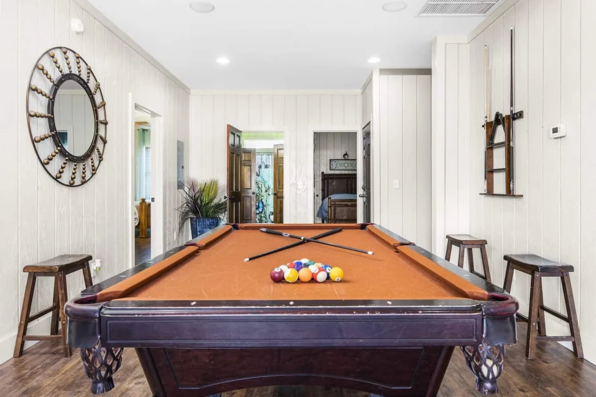 The image showcases a vibrant recreation room featuring a sleek pool table, stylish wooden stools, and a large decorative mirror, all set in a bright, inviting space ideal for group gatherings. The layout encourages socializing and friendly competition, perfect for creating lasting vacation memories.